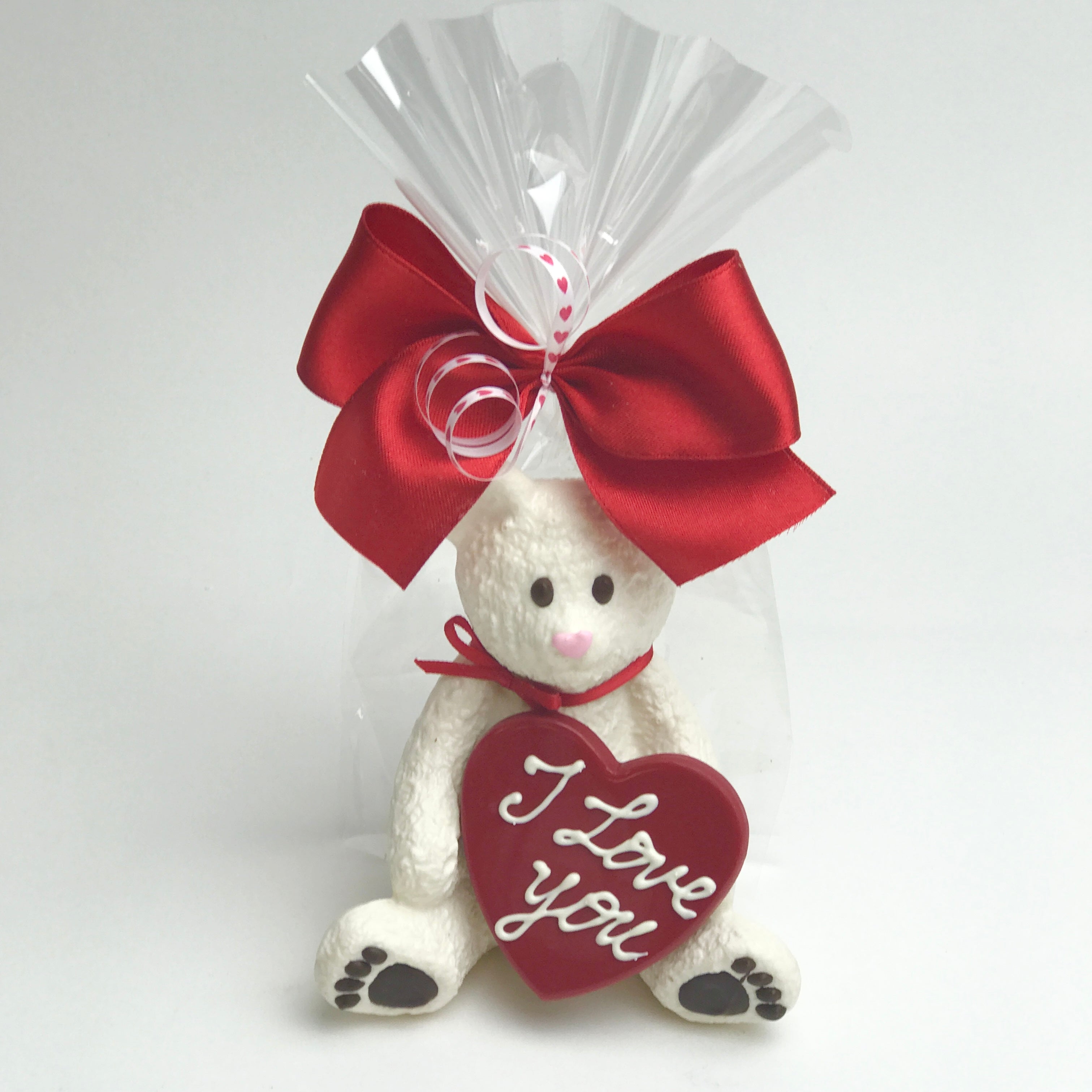 A solid white chocolate bear holding a red heart with "I love you" written on it. The bear is wrapped in clear cello and tied with red and white satin bows.