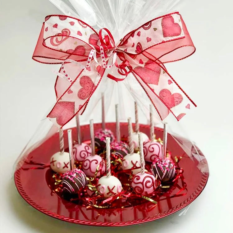 A platter of 12 Valentine's Cake Pop in an assortment of brown, white, and pink designs, wrapped with clear cellophane and a Valentine's bow with red hearts.