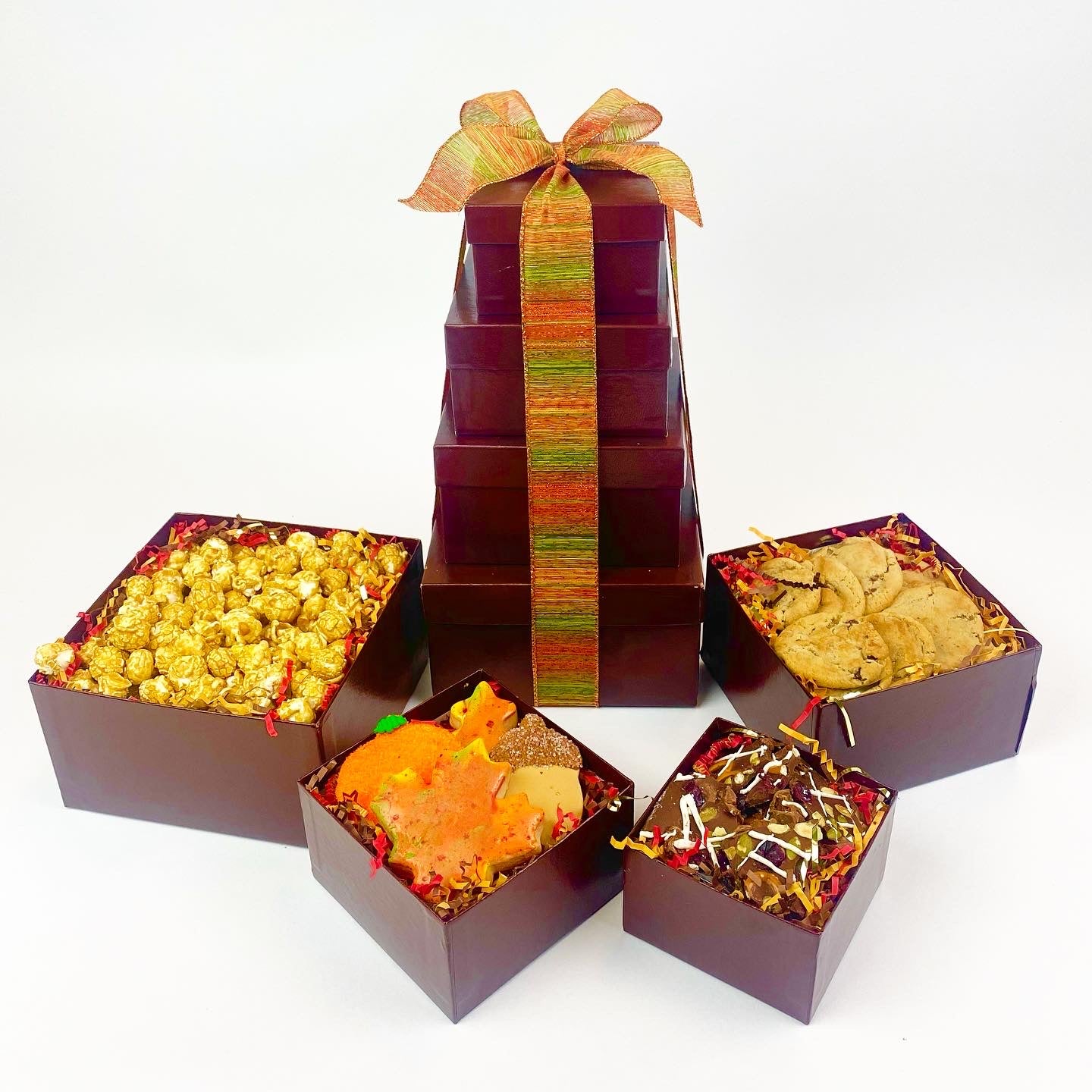 4 brown gift boxes stacked with a gold ribbon. Boxes are filled with chocolate chip cookies, caramel popcorn, decorated sugar cookies and our fall bark