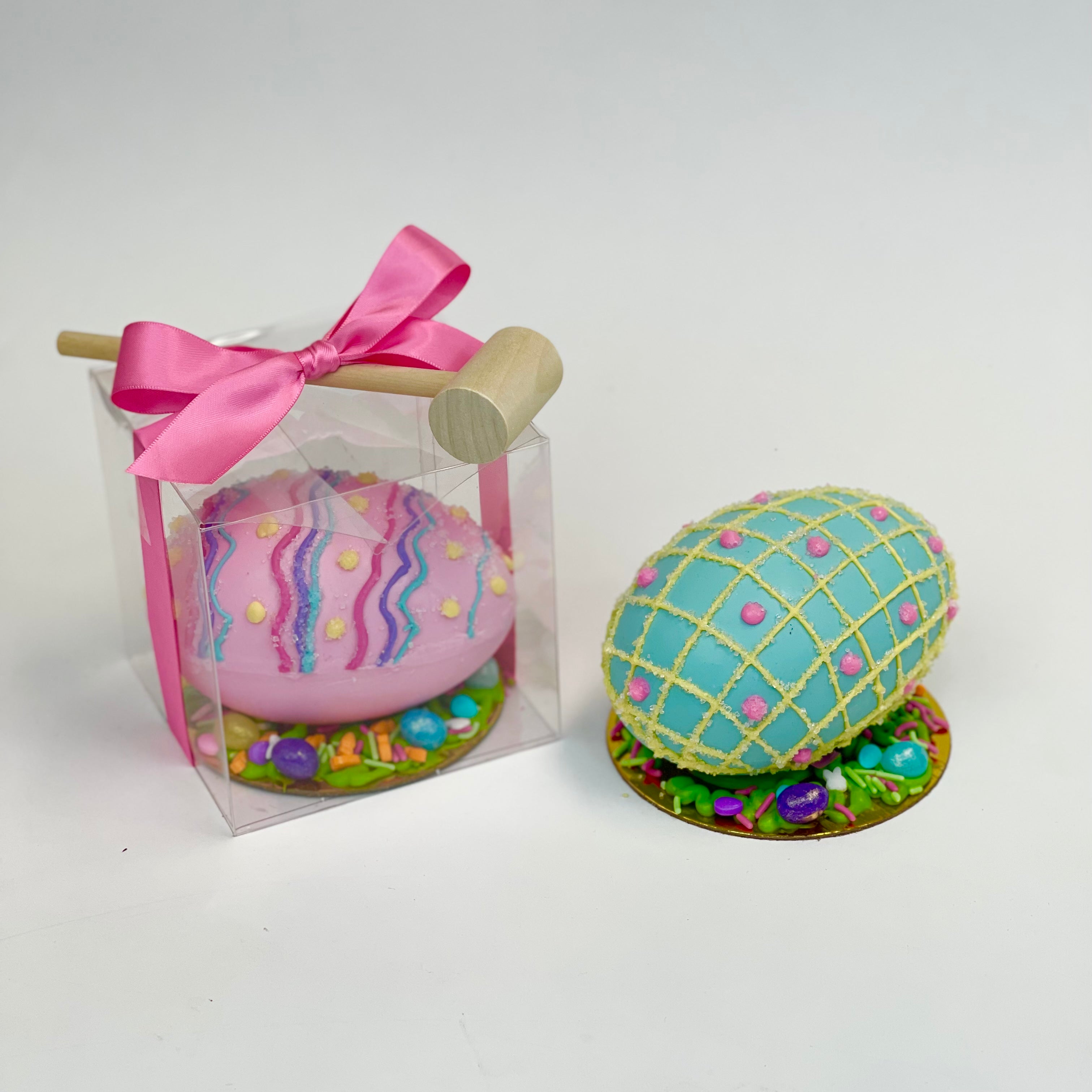 A decorated pink piñata Easter egg inside a clear box, wrapped with a pink ribbon and accompanied by a mallet. Next to it, a decorated blue piñata Easter egg sits on a gold base.