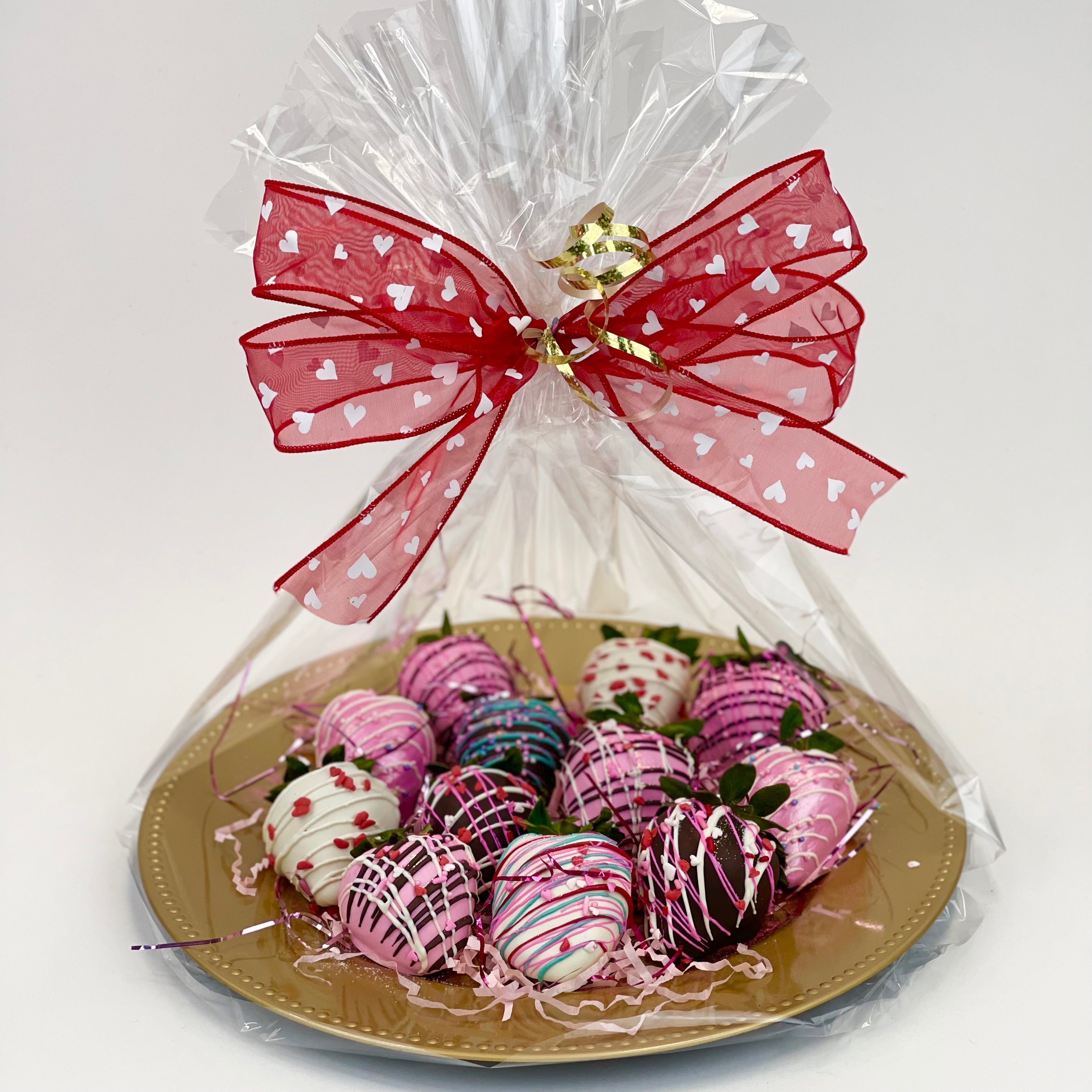 A dozen assorted brown, pink, and white chocolate dipped strawberries beautifully wrapped in clear cello on a golden platter and tied with a big red bow with white hearts.