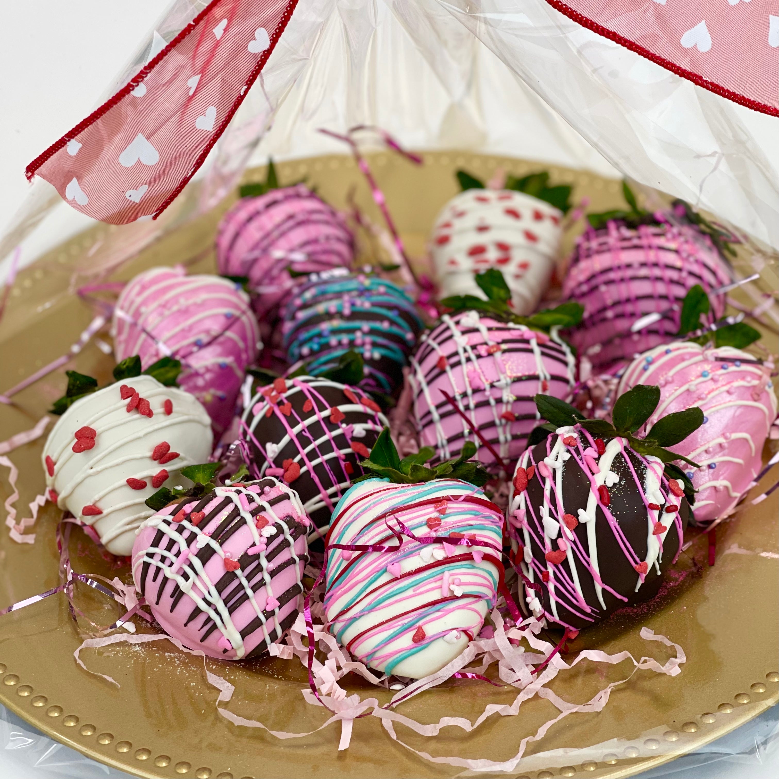 Closeup of a dozen assorted brown, pink, and white chocolate dipped strawberries beautifully wrapped in clear cello on a golden platter and tied with a big red bow with white hearts.