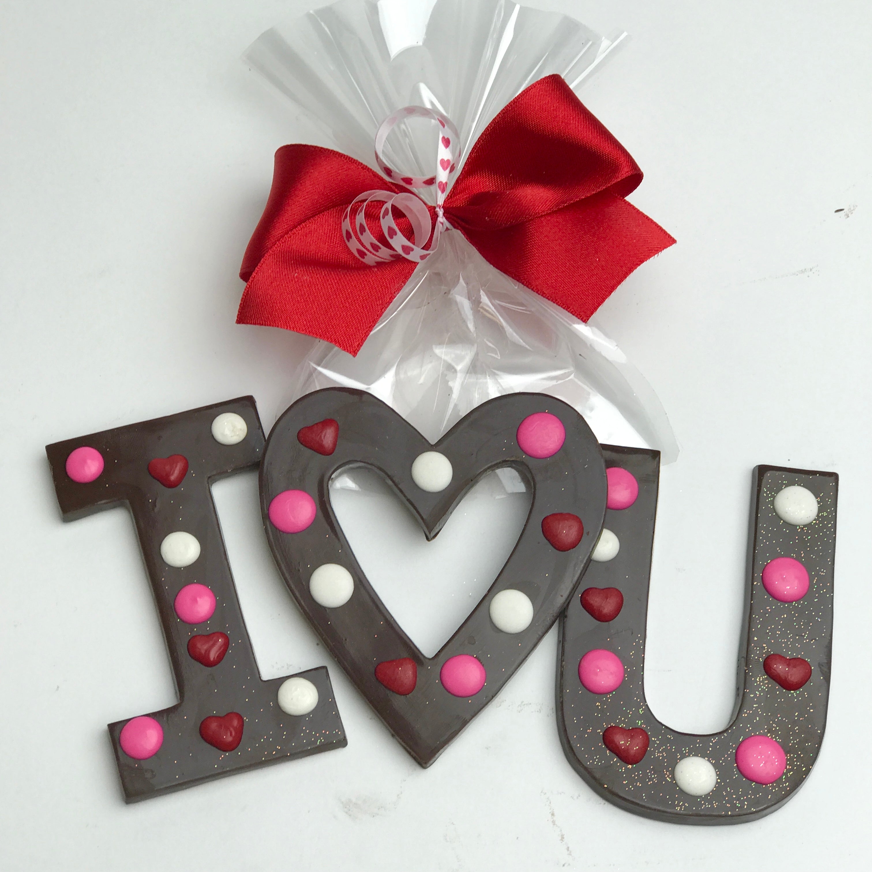 Chocolate letters "I", a heart, and "U" are decorated with red, pink and white dots and wrapped in clear cellophane and tied with red and white satin bows.