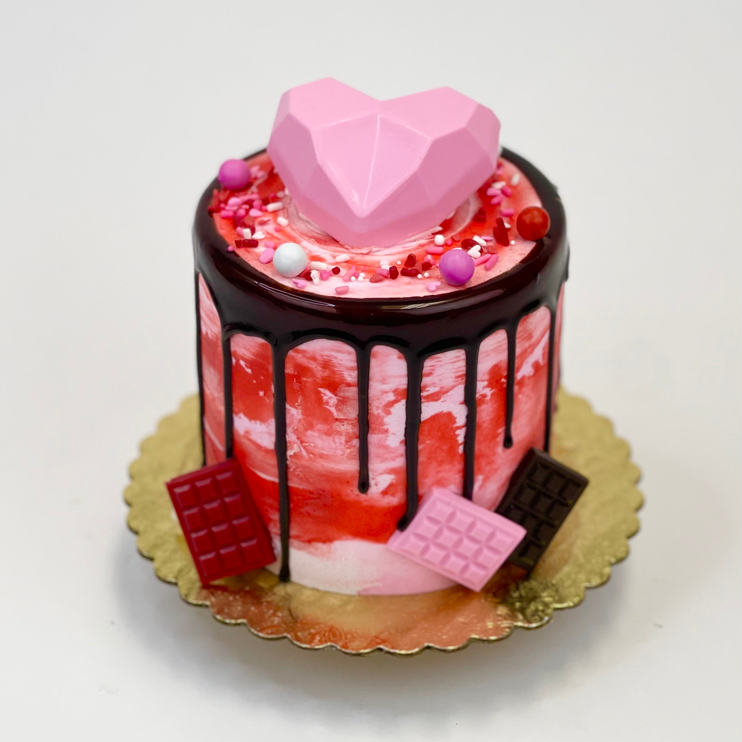 iced in pink and red buttercream with pink chocolate geode heart and sixlet candies on top. Drizzled with chocolate ganache and 3 small chocolate bars resting on board in pink, red and chocolate. Sits on a gold board