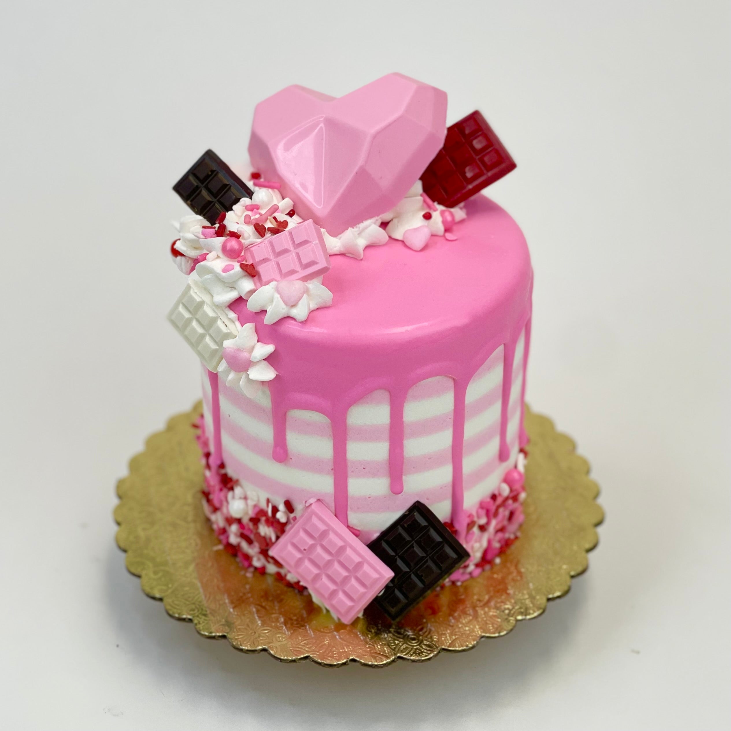 A 4” cake iced in pink and striped buttercream design with chocolate hearts and small chocolate bars on top on a pink chocolate drip.