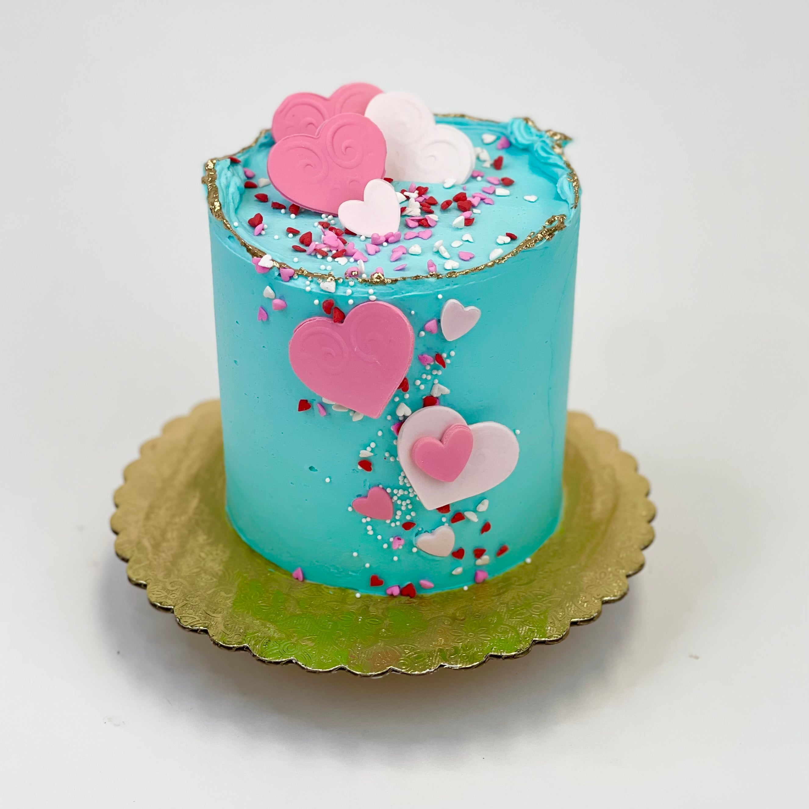 4” cake iced in a pretty tiffany buttercream design with fondant hearts in shades of pink and matching sprinkles then brushed with a light gold accent. Cake sits on a gold board