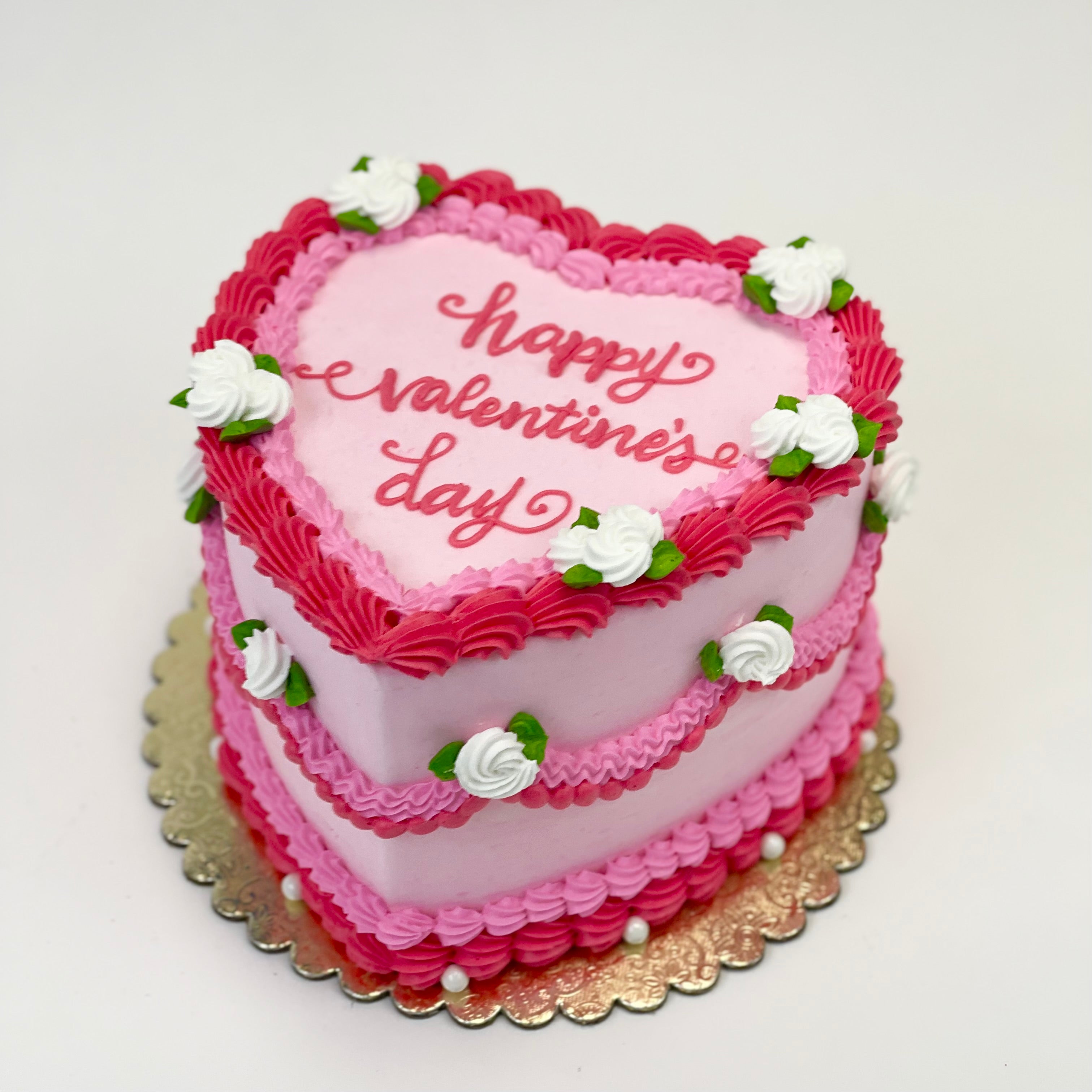 A 6" heart cake iced in light pink with red and hot pink accents, adorned with white flower rosettes and green leaves.